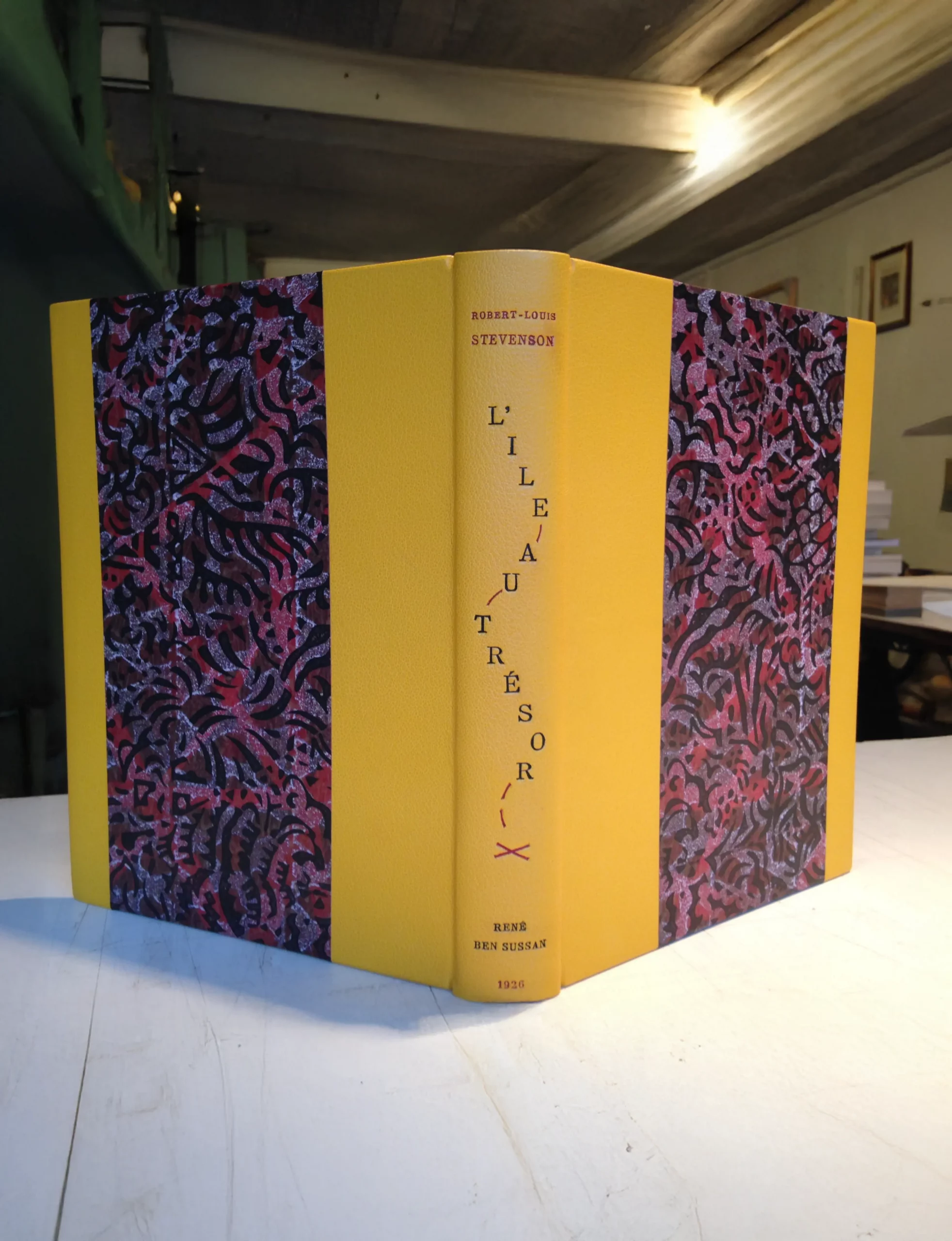 Reliure artisanale de L’Île au trésor de Robert-Louis Stevenson, demi-cuir jaune avec motifs décoratifs rouges et noirs, réalisée par l’Atelier Reliure Paris